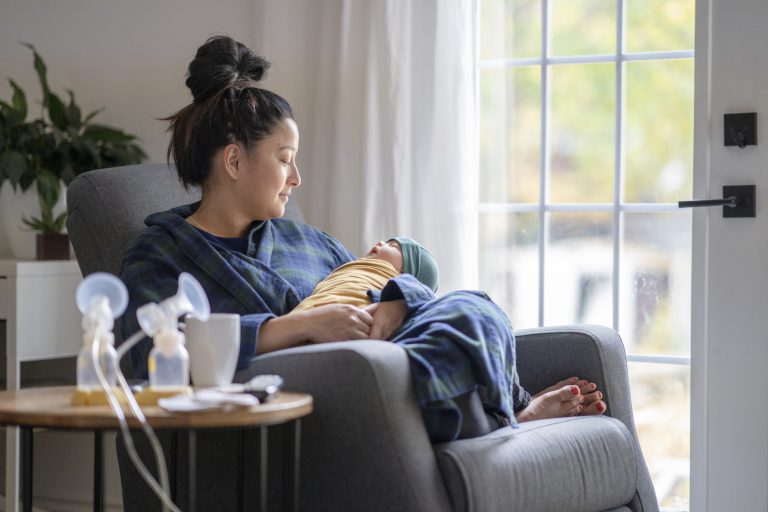 Parent sits in chair holding newborn with breast pumps on table next to them.