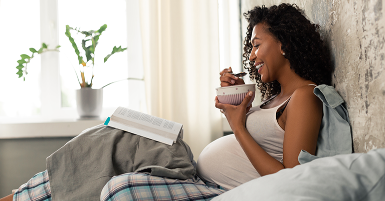Pregnant woman eating breakfast in bed while reading.