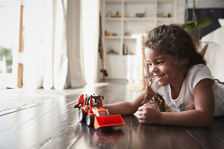 Young child with long hair plays with toy truck while laying on the floor.