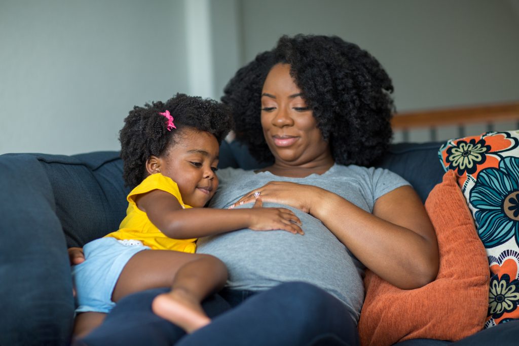 Black pregnant woman laying on couch with toddler daughter and looking at pregnant belly together.