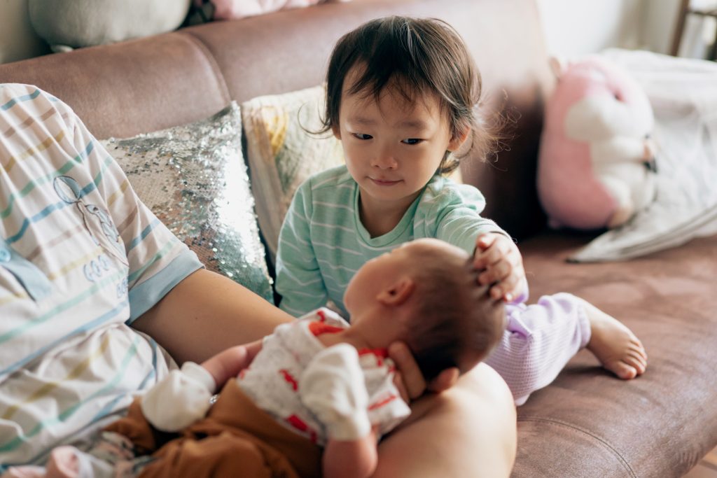 Asian toddler sitting on couch next to parent while stroking hair of newborn sibling.