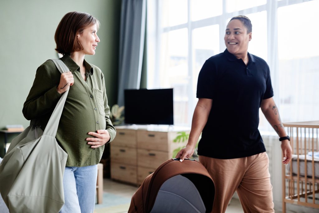 LGBTQIA+ couple with pregnant person walking out of room with partner that is holding a baby carrier.