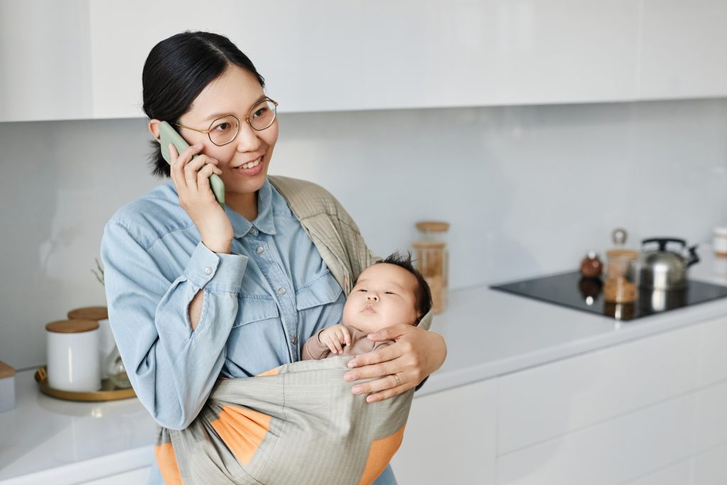 young mom holding baby in sling while having conversation on mobile phone