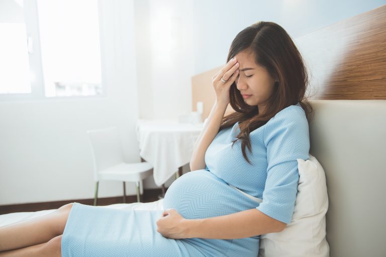 A portrait of a pregnant woman has headache sitting on her bed