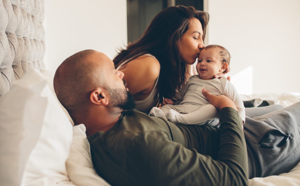Parents with their newborn baby boy on bed at home. Woman kissing her son sitting with father.