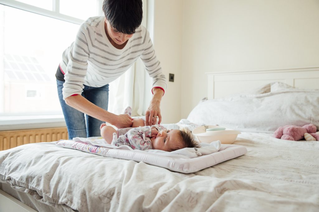 Baby has had her nappy changed by her mother on the double bed. The mother is fastening the baby grow.