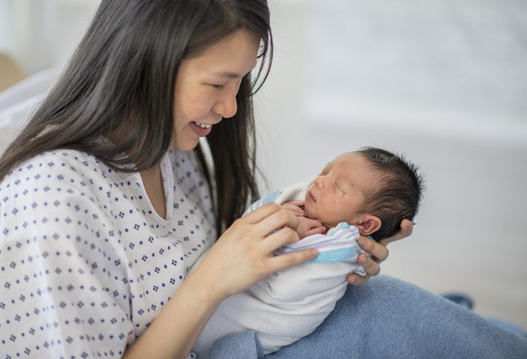 Una madre con bata de hospital sonríe con ternura a su recién nacido, quien está envuelto en una manta blanca con rayas azules.