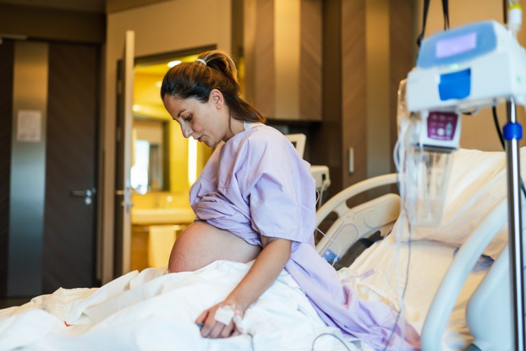 Mujer embarazada con bata de hospital sentada en una cama, mirando hacia abajo, con una vía intravenosa conectada.