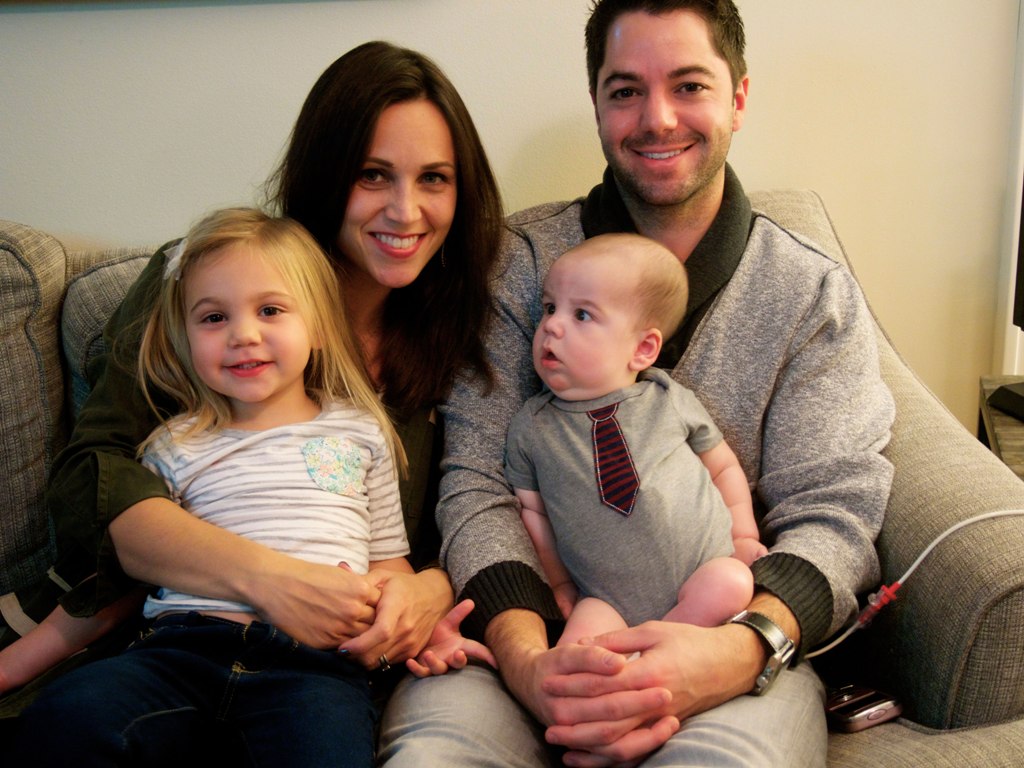 Lindsey and Eddie sit on a couch at home, holding their two children Lucy and Rocco.