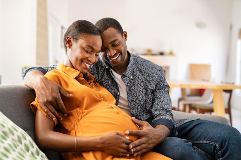 Una pareja sonriente está sentada en un sofá, con el hombre abrazando cariñosamente a la mujer, que está embarazada. Ambos están tocando su vientre con ternura.