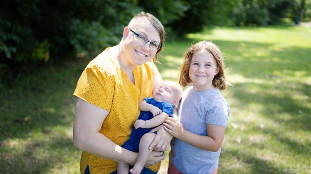 smiling woman holding an infant with child standing next to her